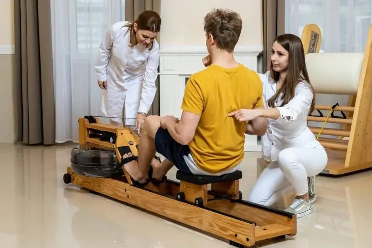 Páciens evezőgépen ül, két gyógytornász figyeli és korrigálja a mozgását