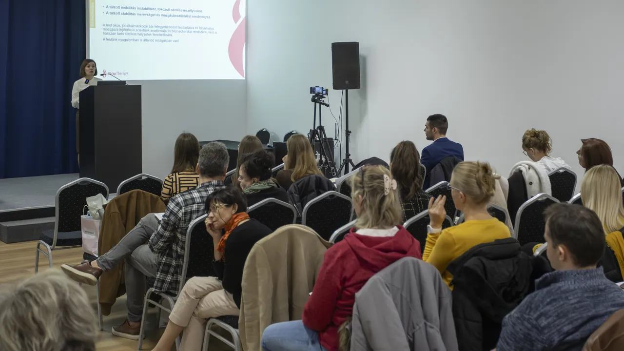 Közönség hátulról egy előadóteremben, elöl előadó és vetítés